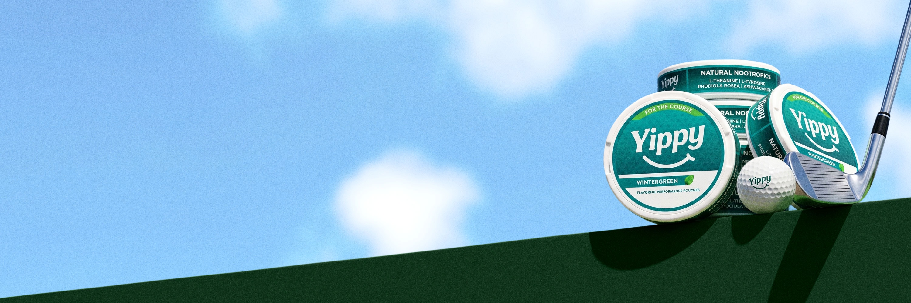 Still life of Yippy For The Course Wintergreen tins on a green putting surface with a golf ball and putter under a blue sky with drifting clouds