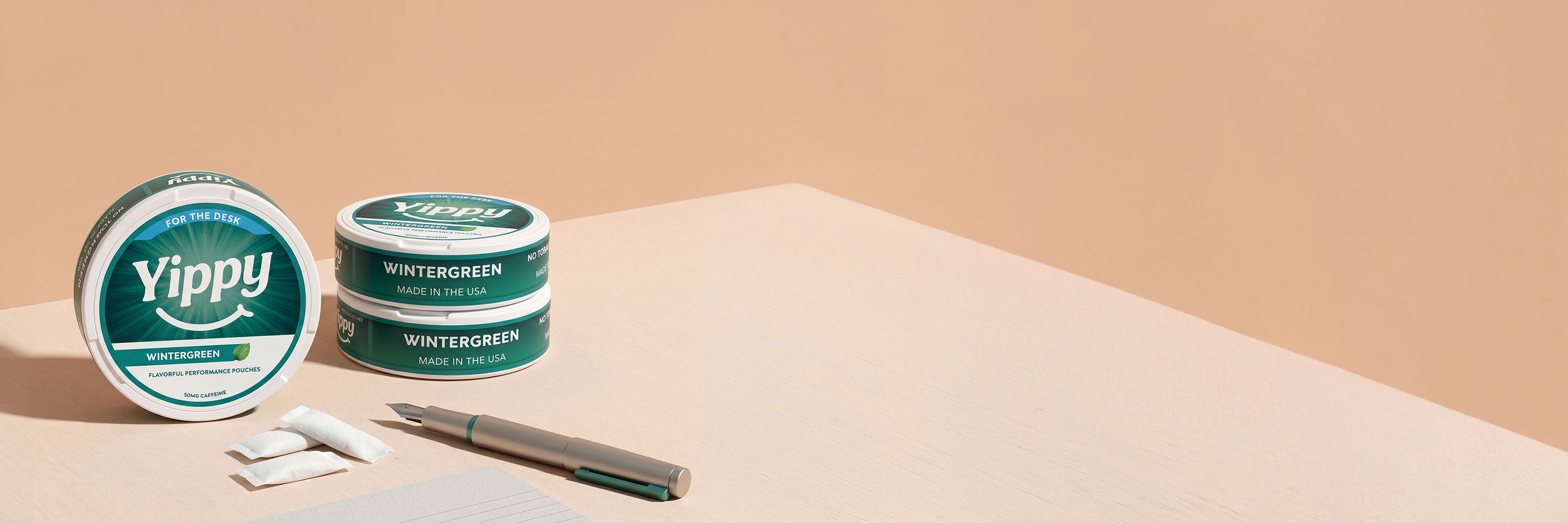 Still life of Yippy For The Desk Wintergreen tins arranged with a fountain pen, paper, and pouch packets on a peach paper backdrop